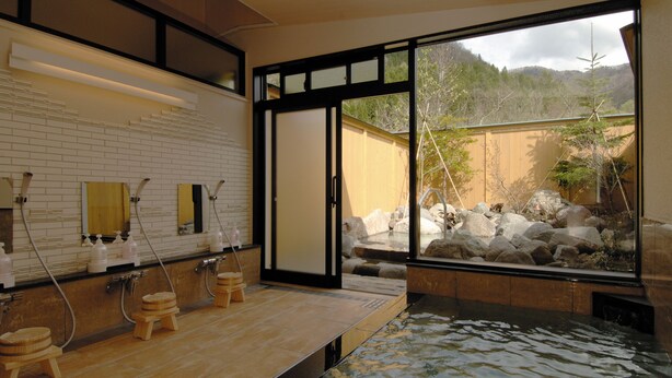 A traditional Japanese onsen with a wooden platform, a stone bath, and a view of a mountainous landscape through large windows. - Oyado Nozawa (Takayama)