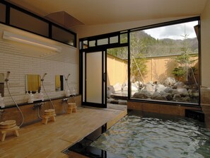 Una tradizionale onsen giapponese con una piattaforma in legno, una vasca in pietra e vista su un paesaggio montuoso attraverso ampie finestre.