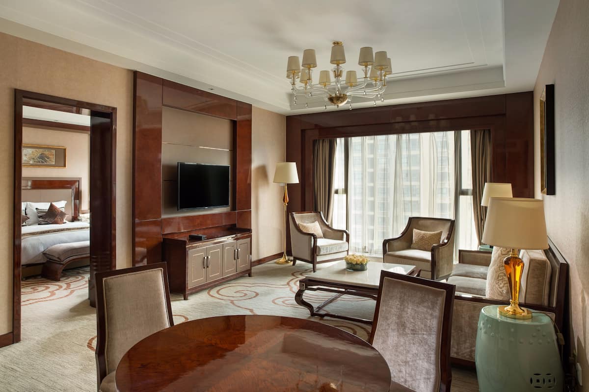 Una habitación de hotel con una cama grande, televisión, comedor con mesa y sillas, y zona de estar con sillones y una mesita de centro.