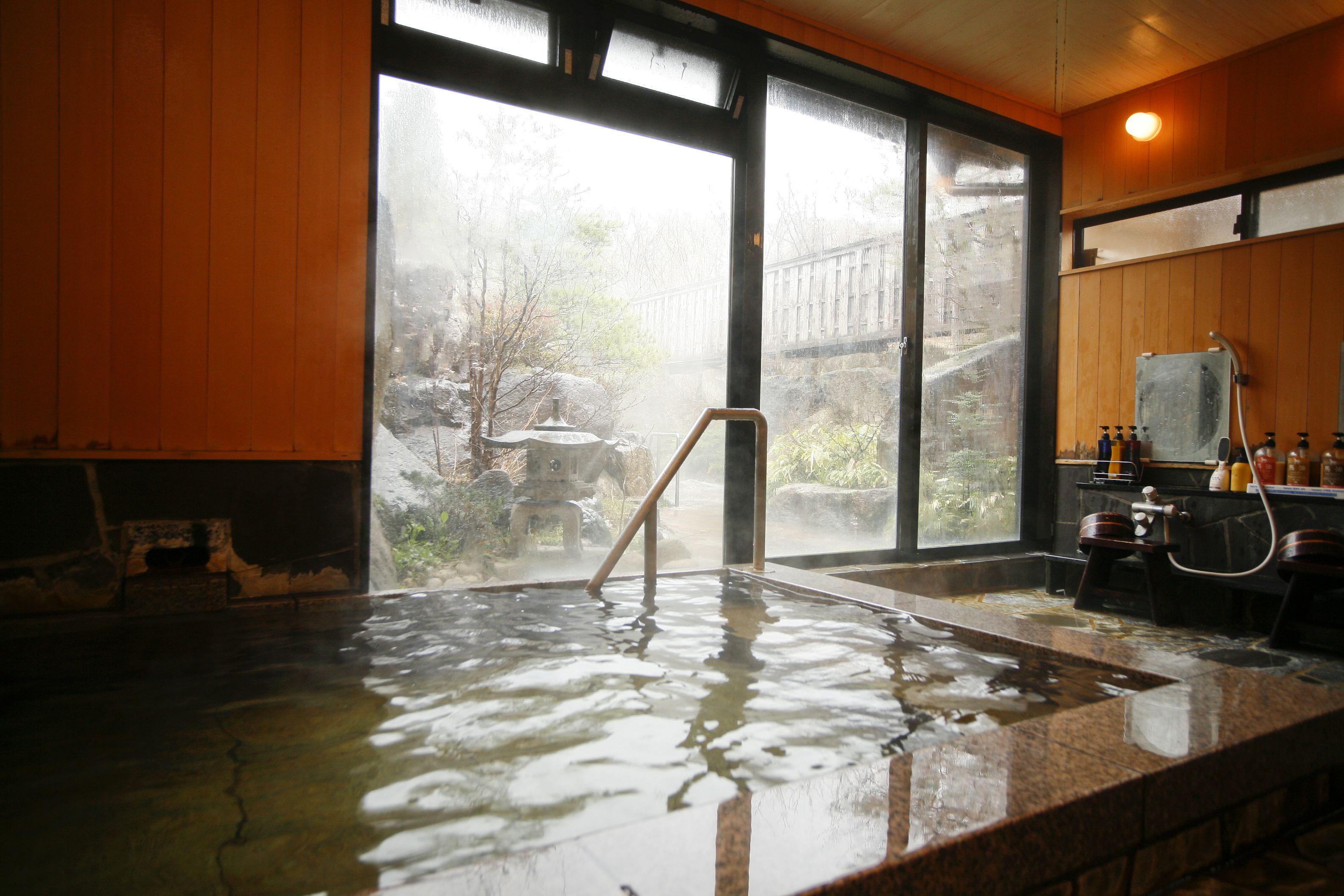 A hot spring bath with a wooden interior, a view of a forest through large windows, and a wooden ceiling.