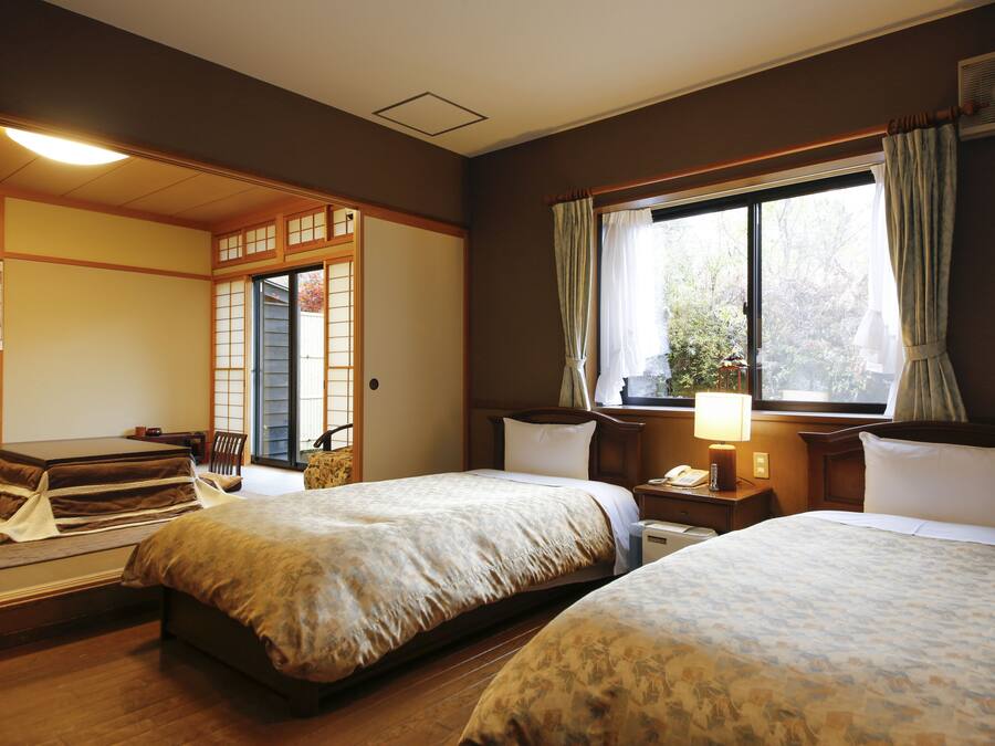 A traditional Japanese-style hotel room with tatami flooring, two beds, a sliding door, and a view of the outdoors.