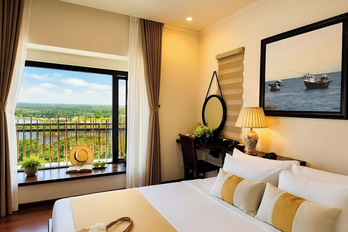 A hotel room with a bed, a desk with a lamp, a window with a view of the sea, and a painting on the wall.