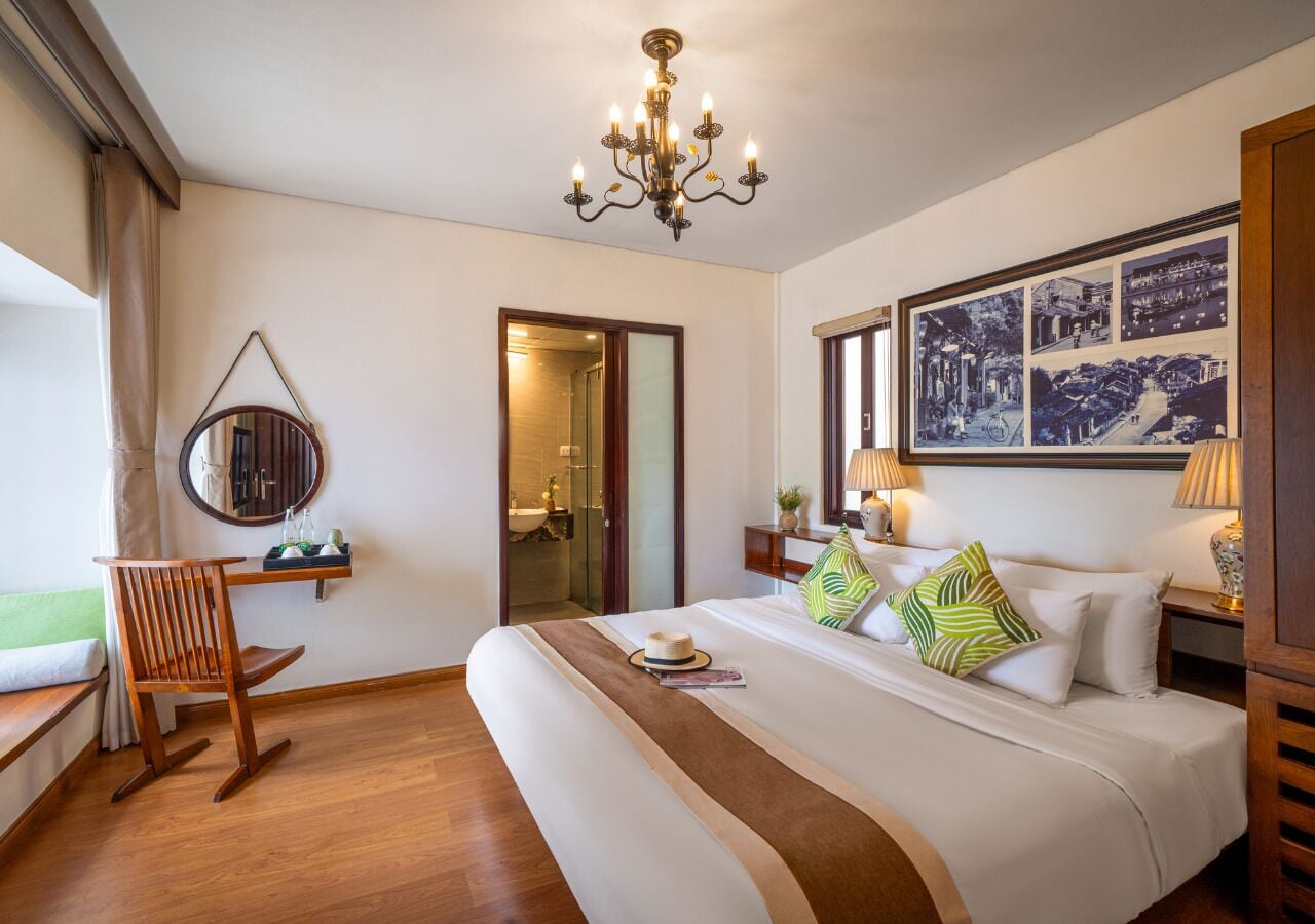 A hotel room with a large bed, a wooden rocking chair, a chandelier, and a framed picture on the wall.