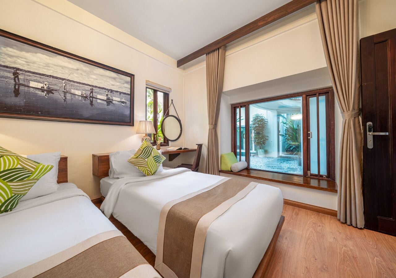 A hotel room with two beds, a desk, a window with a view of the outdoors, and a painting on the wall.