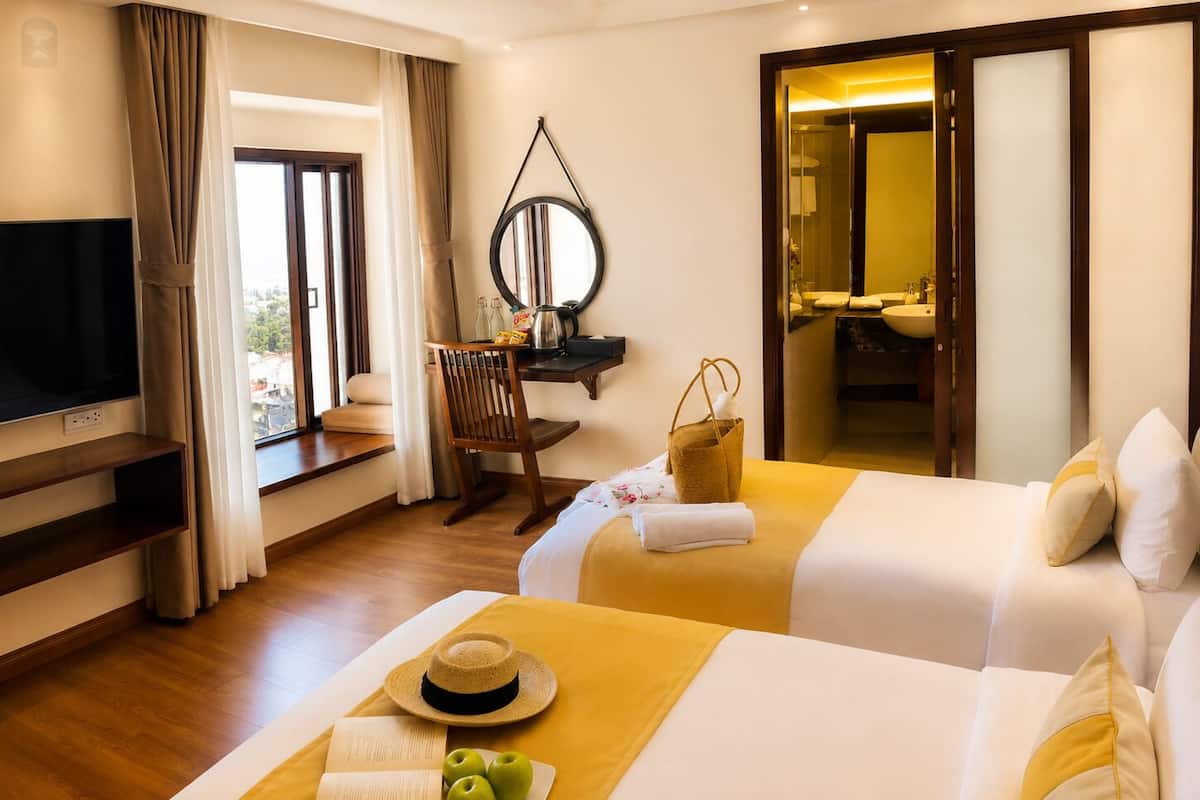 Habitación de hotel con dos camas, televisor, escritorio con silla, espejo y baño visible a través de una puerta abierta.