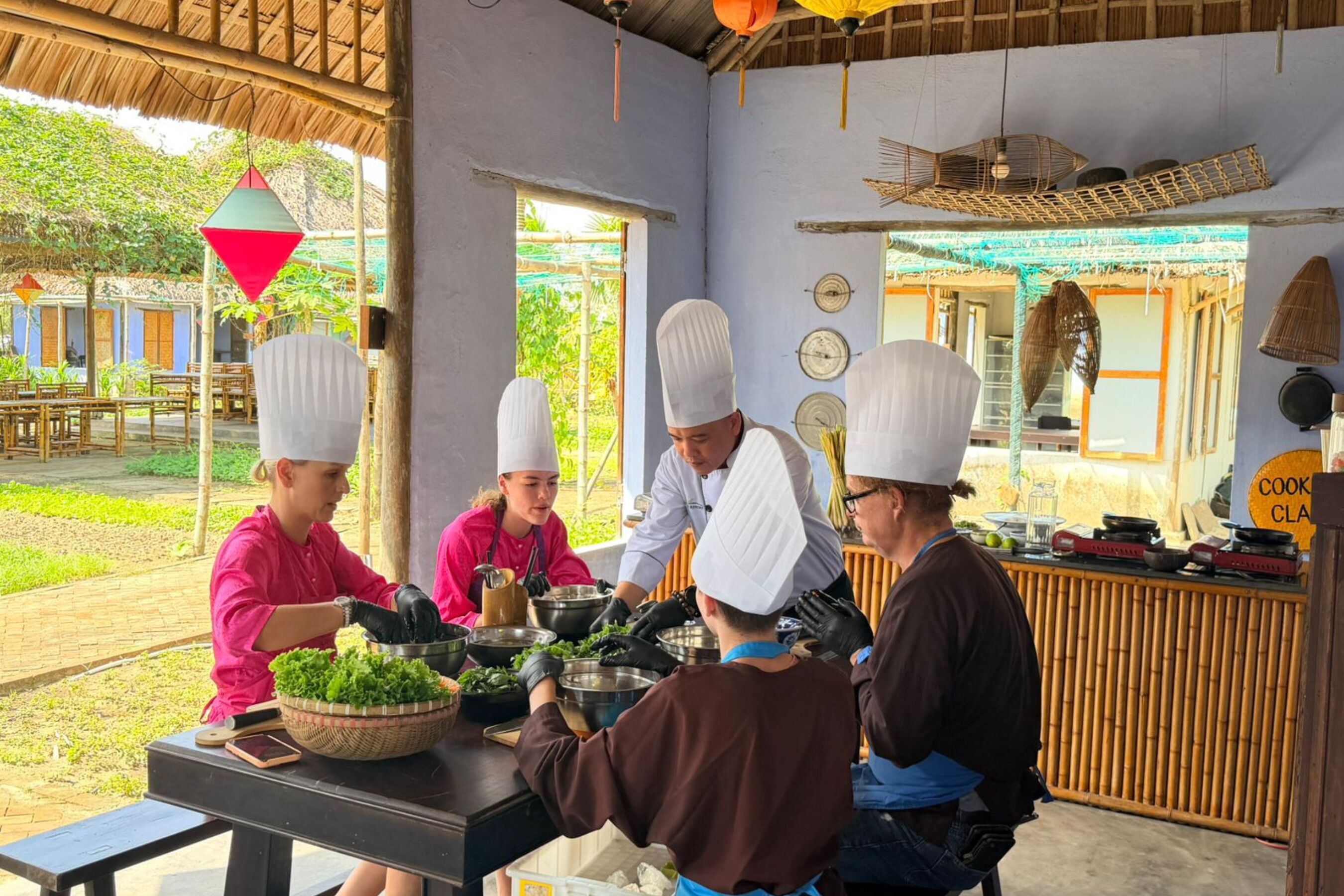 A group of chefs preparing food in a rustic outdoor kitchen with bamboo elements and a thatched roof.