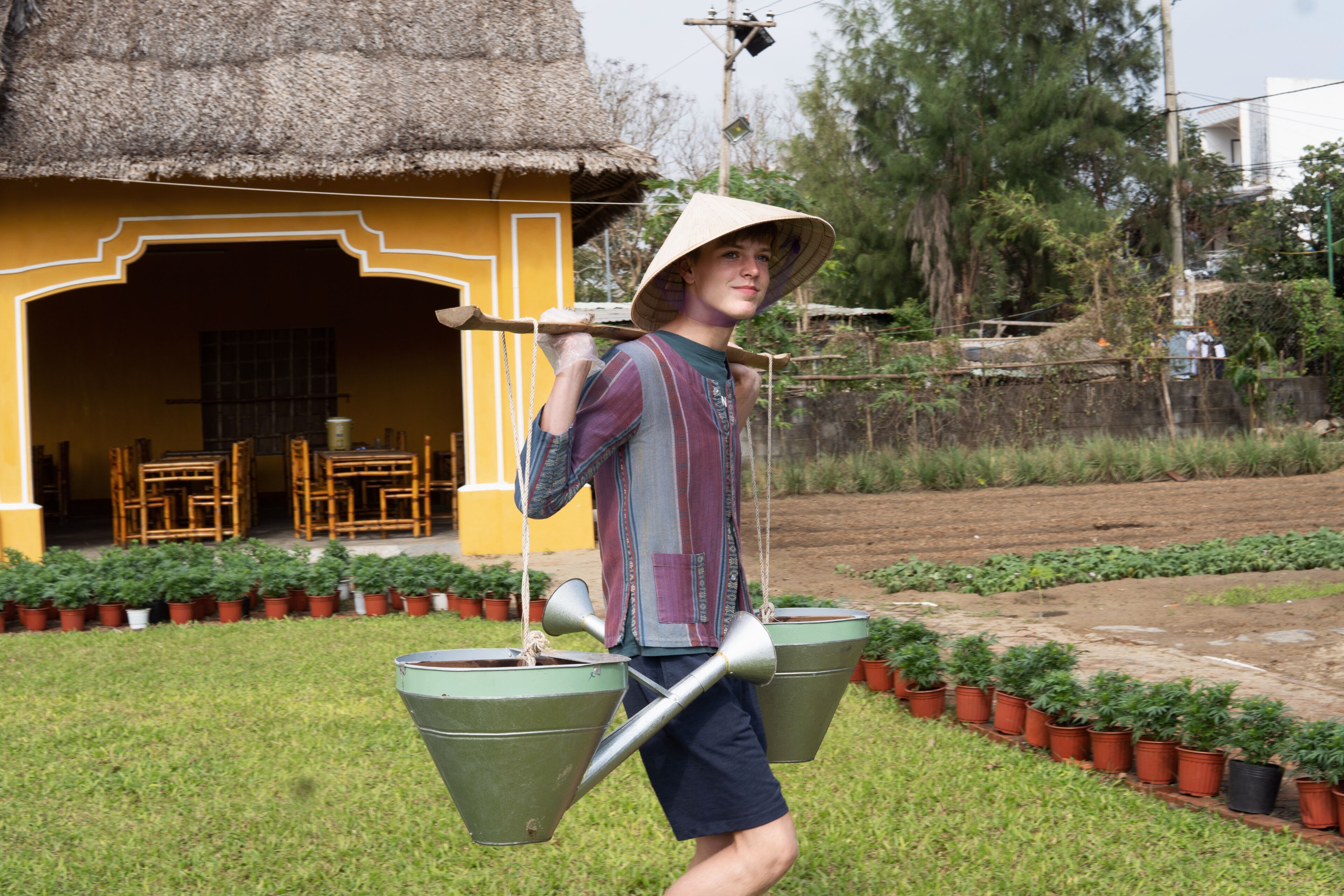 A person carrying a large metal watering can and a smaller one, standing in front of a yellow building with a thatched roof.
