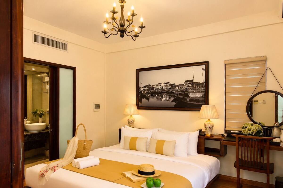 Habitación de hotel con cama, lámparas de noche, un cuadro enmarcado, un espejo y un baño visible a través de una puerta abierta.