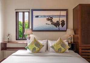A hotel room with a bed, two lamps, a window with a view of boats, and a painting on the wall.