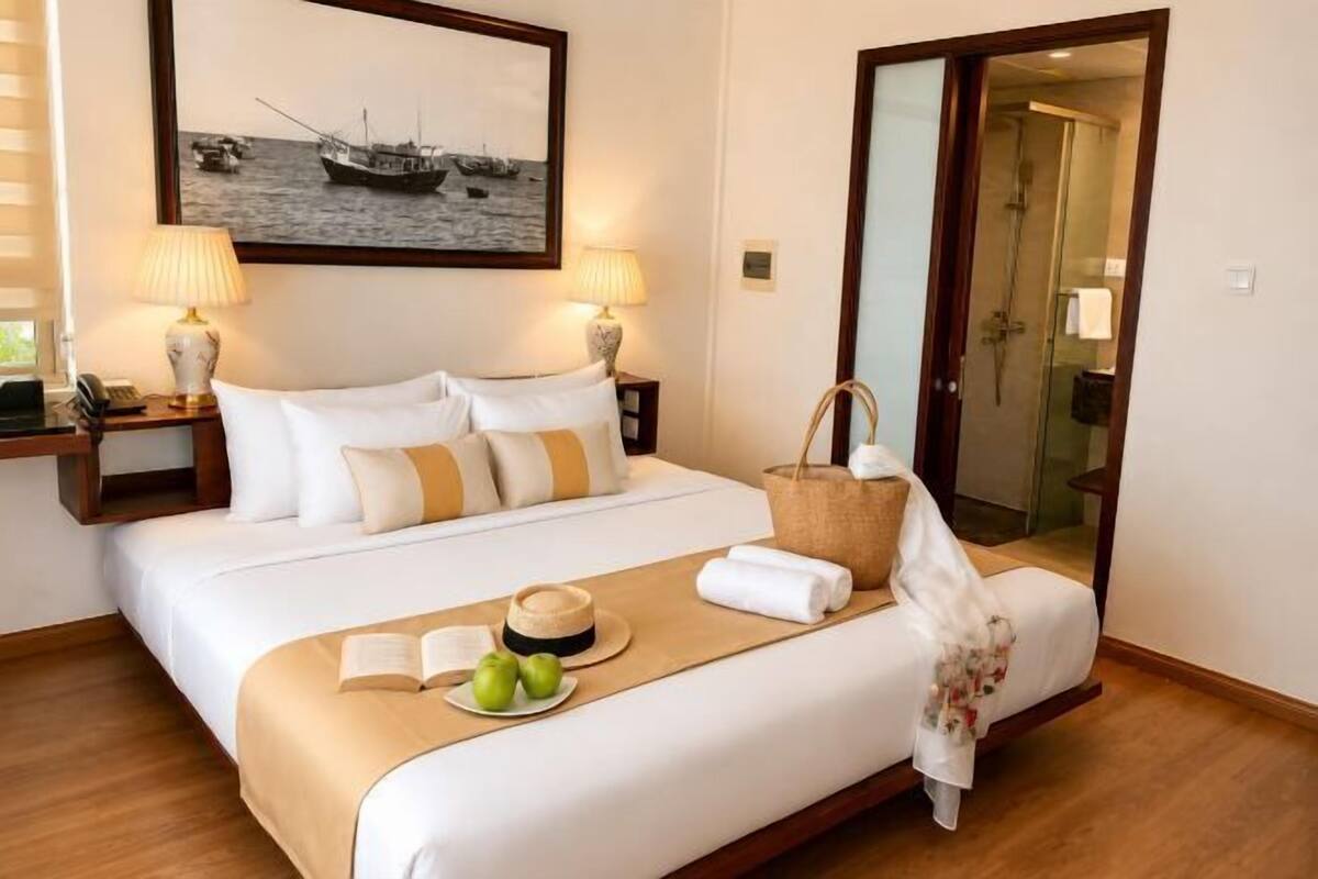 Una habitación de hotel con cama, un cuadro en la pared, una mesita de noche con lámpara y un baño visible a través de una puerta de vidrio.