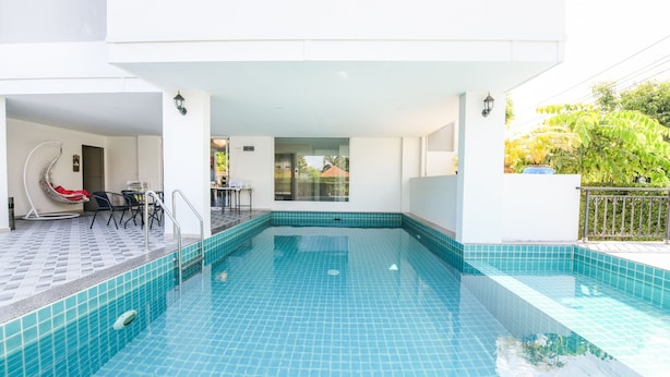 Une piscine dont les bords sont recouverts de carreaux, un espace de repos couvert avec des chaises, et une vue sur un bâtiment en arrière-plan.
