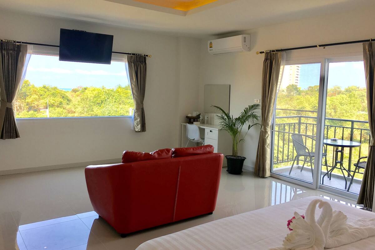 Une chambre d’hôtel avec un canapé rouge, un lit avec une literie blanche, une plante en pot et un balcon agrémenté de mobilier d’extérieur.