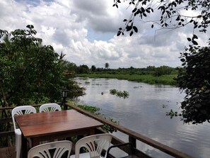 A wooden table and white chairs by a calm river, surrounded by lush greenery and a cloudy sky. - Be Fine Sabuy Hotel & Resort (Surat Thani)