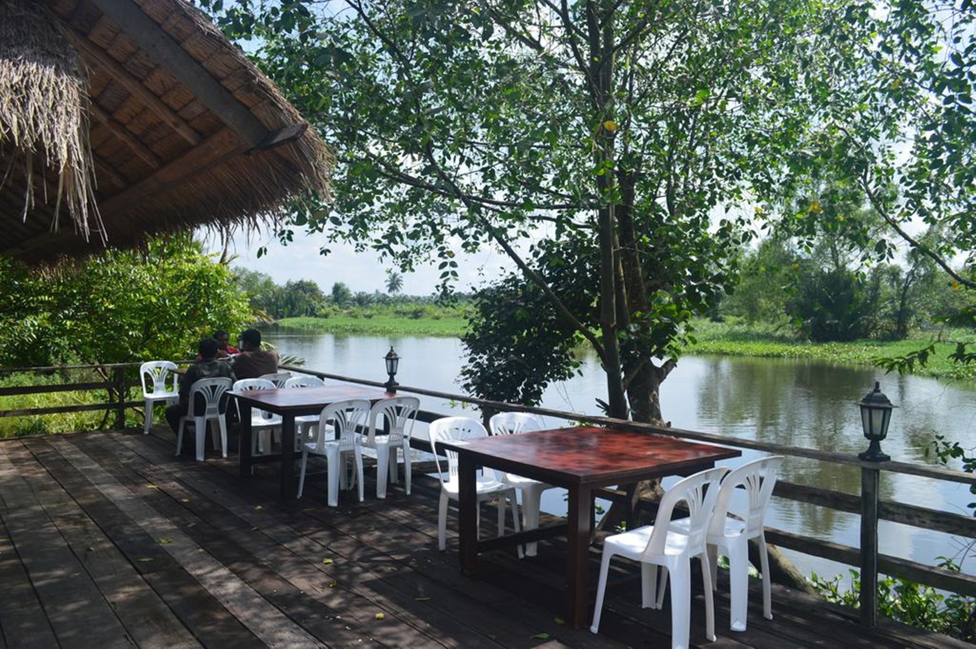 Un espace de restauration en plein air, avec des tables et des chaises, un toit de chaume et une vue sur un lac entouré de verdure.