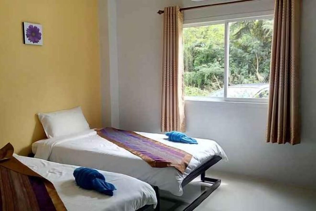 A room with two beds, a window with curtains, and a view of greenery outside.