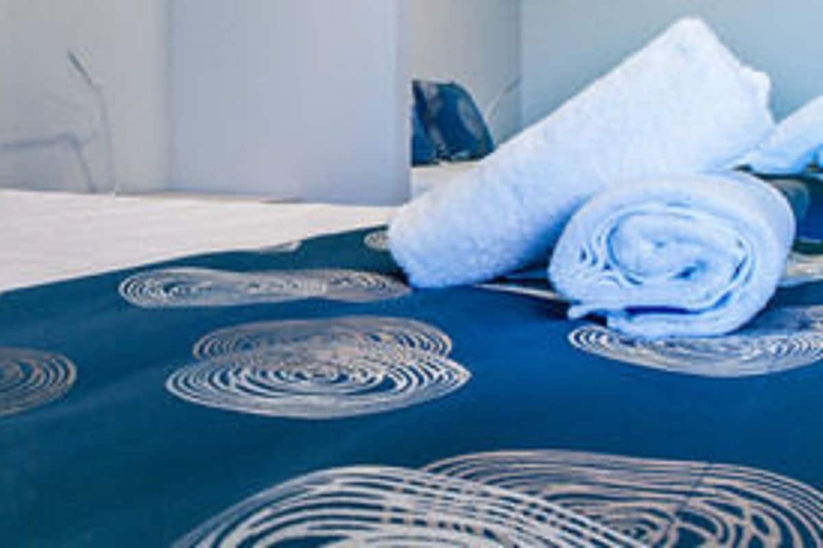 Una cama bien hecha con una colcha azul adornada con diseños circulares, dos almohadas con diseños parecidos y una toalla blanca enrollada sobre la cama.