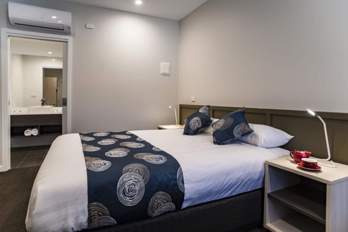 Habitación de hotel con cama, mesita de noche con lámpara, espejo y baño visible a través de una puerta abierta.
