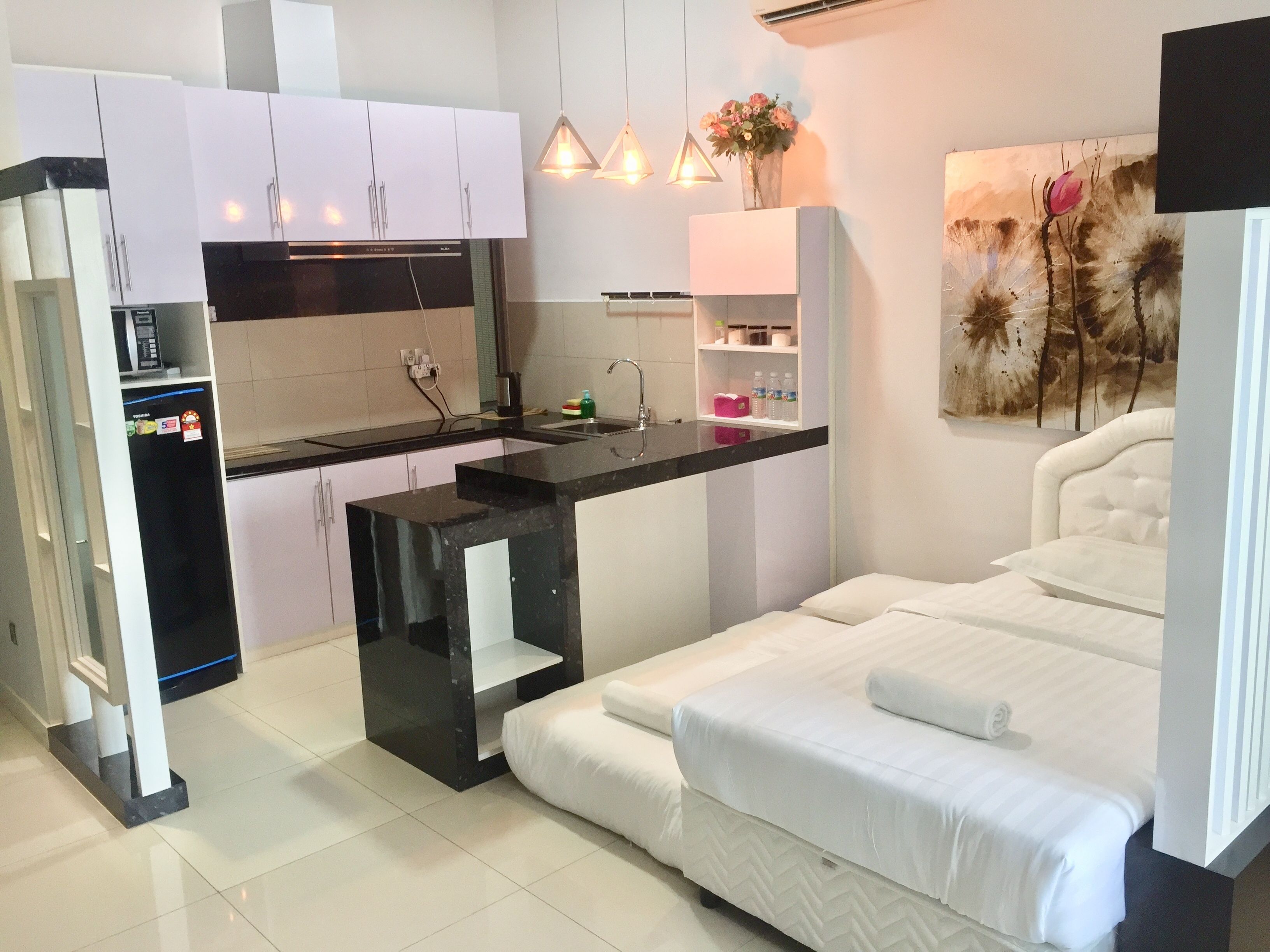 A compact hotel room with a kitchenette, a bed with white bedding, and a painting on the wall.