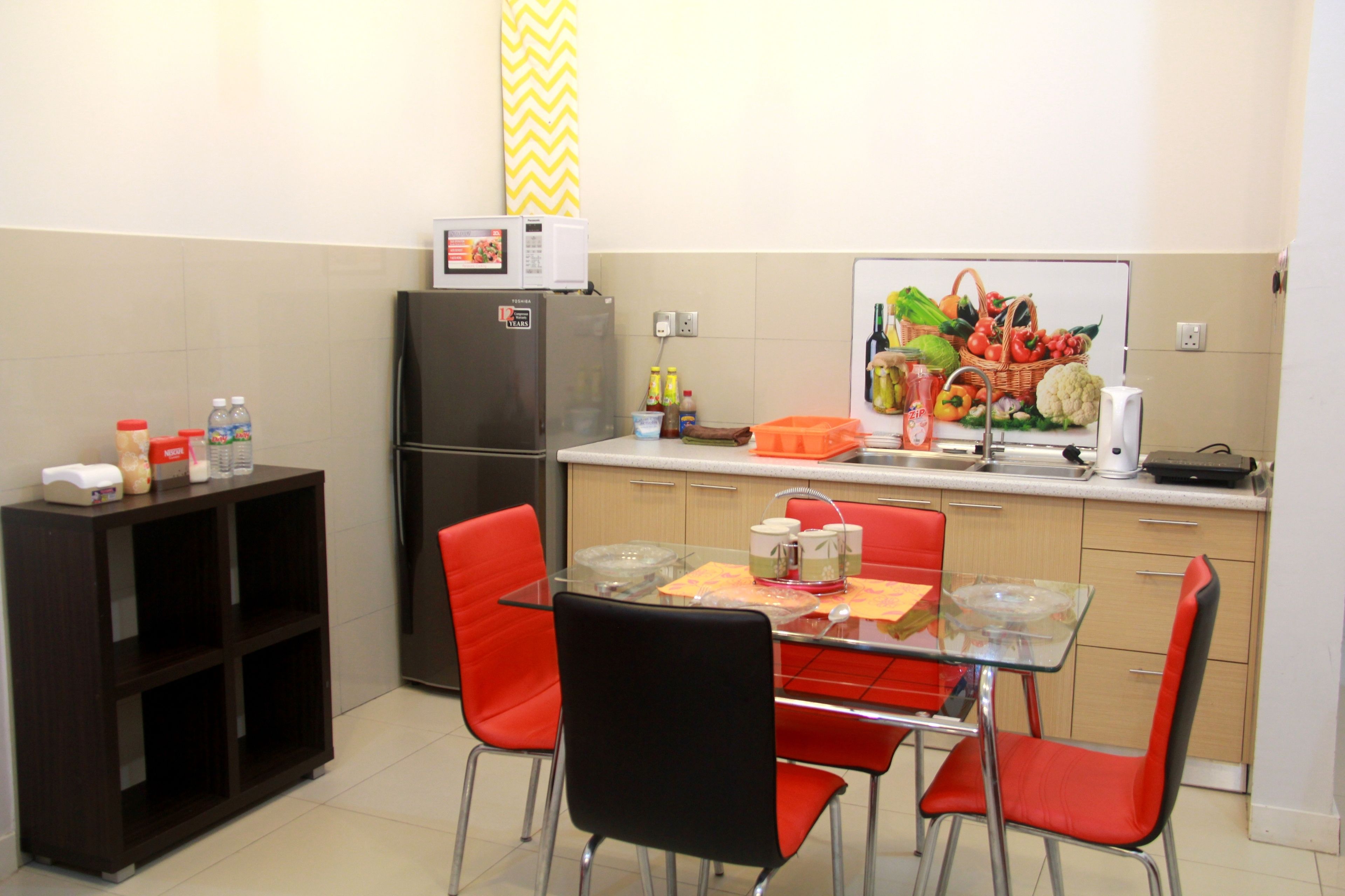 A compact kitchen with a refrigerator, microwave, and a dining area with red chairs and a glass table.