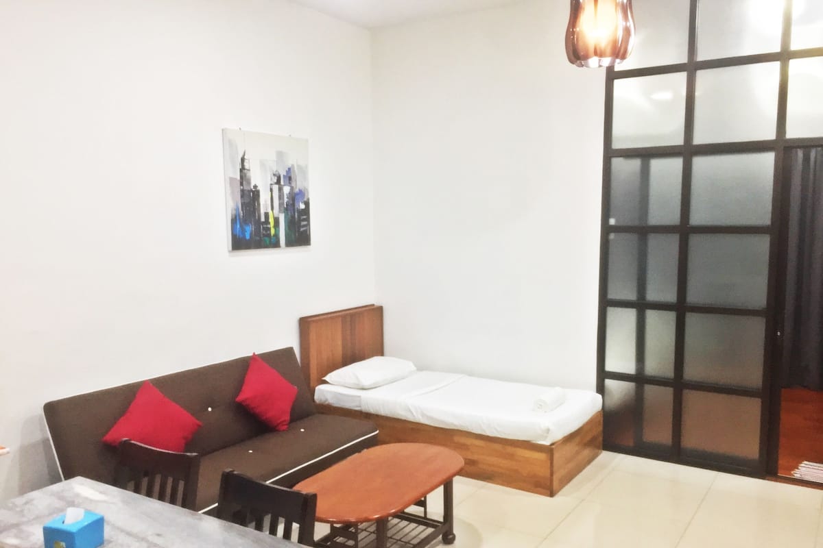 Habitación compacta con cama, sofá con cojines rojos, una mesita y una silla. Hay un cuadro en la pared y una puerta con diseño de cuadrícula.