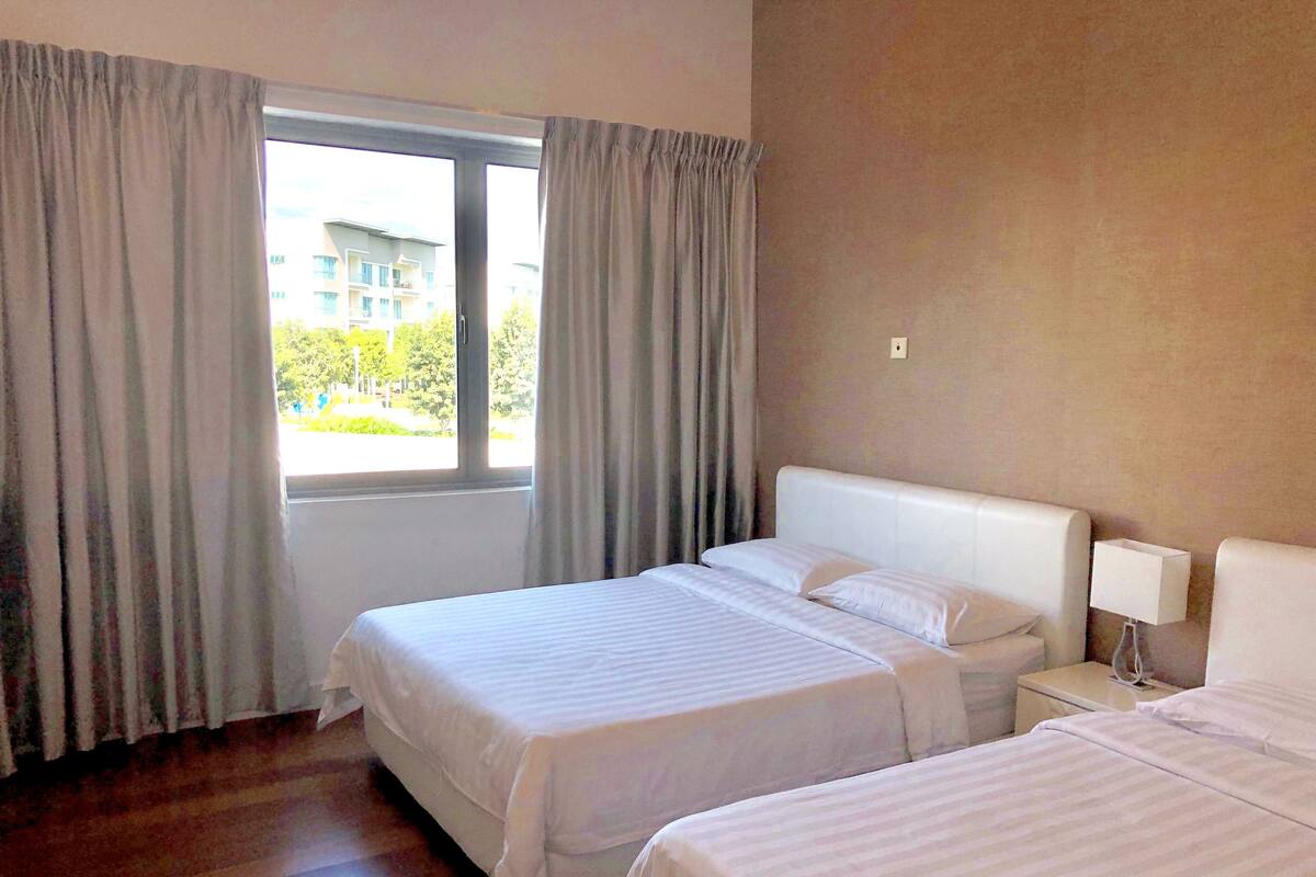 Habitación de hotel con dos camas, una ventana con cortinas, una lámpara de noche y vistas a edificios.