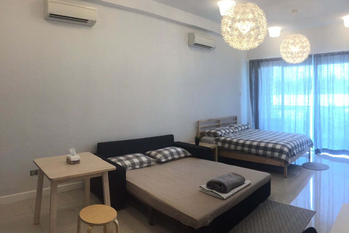 A modern hotel room with a large bed, a smaller bed, a desk, and a chair. There are air conditioning units on the wall, a ceiling light fixture, and a window with curtains.