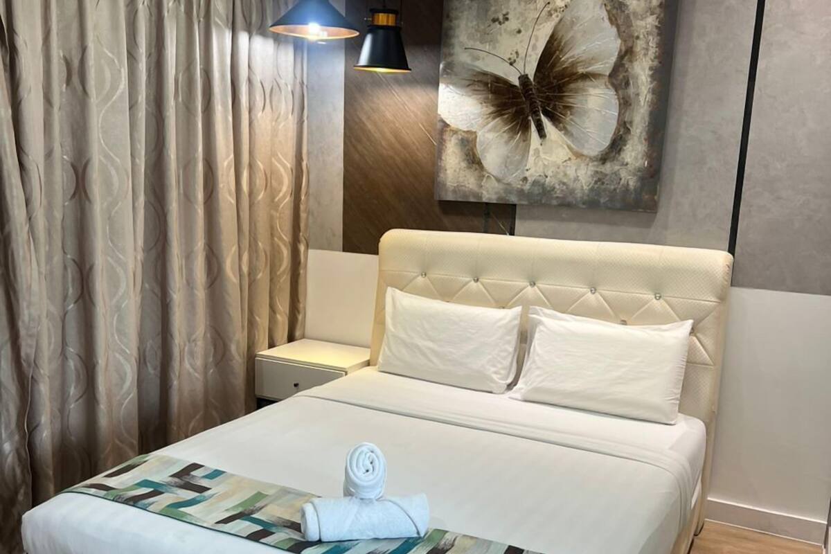 Una cama bien hecha con una toalla enrollada, una obra de arte en la pared de una mariposa y una mesita de noche con una lámpara.