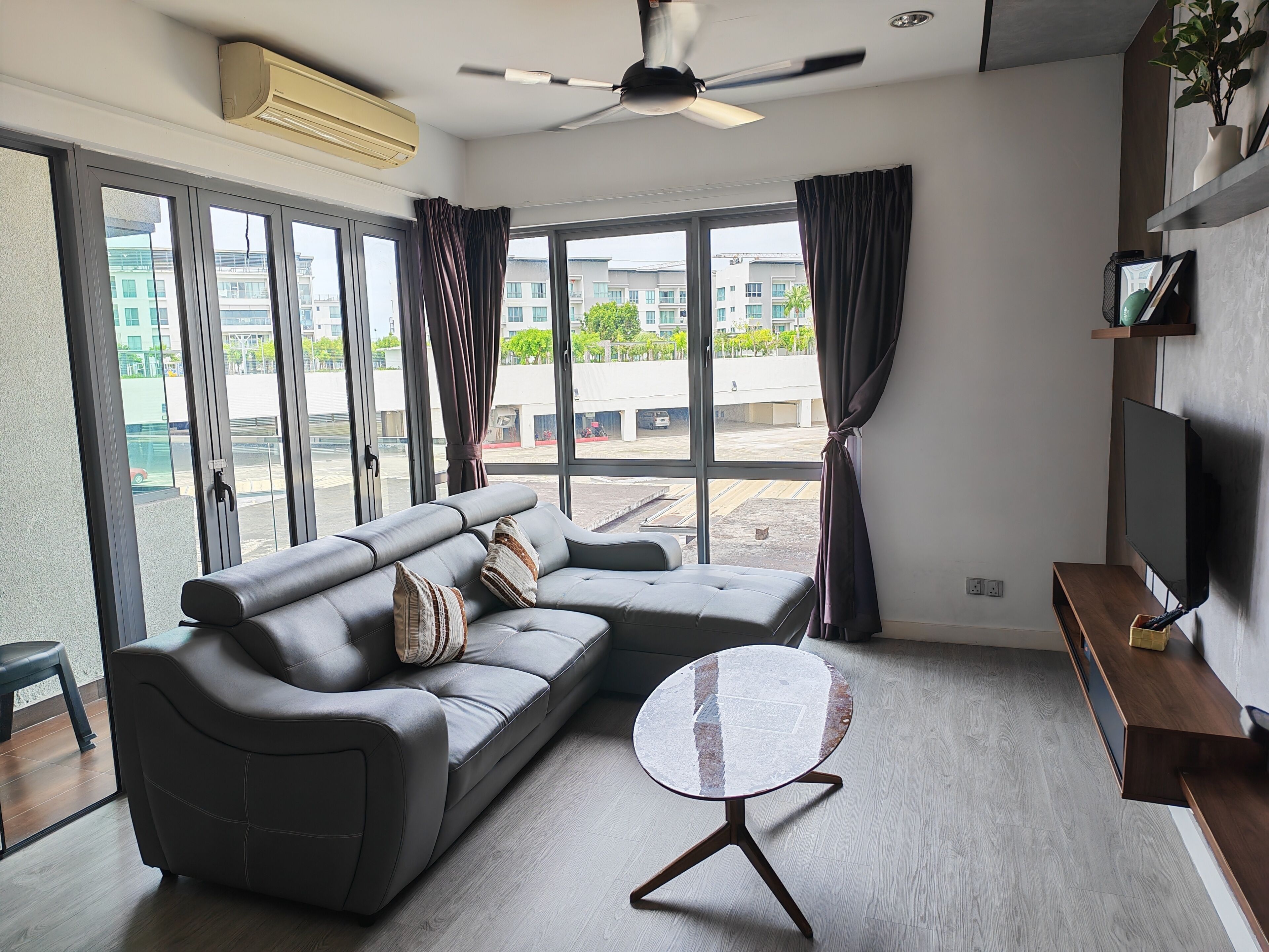 A modern living room with a grey sofa, a round coffee table, a ceiling fan, and a large window with curtains.