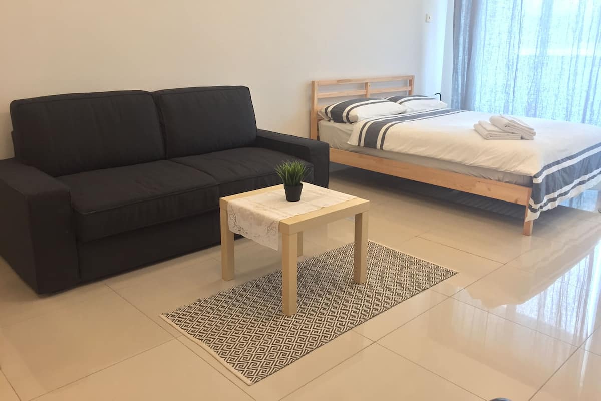 Una habitación moderna con un sofá negro, una mesita con una planta, una cama bien hecha con ropa de cama a rayas y una alfombra estampada.