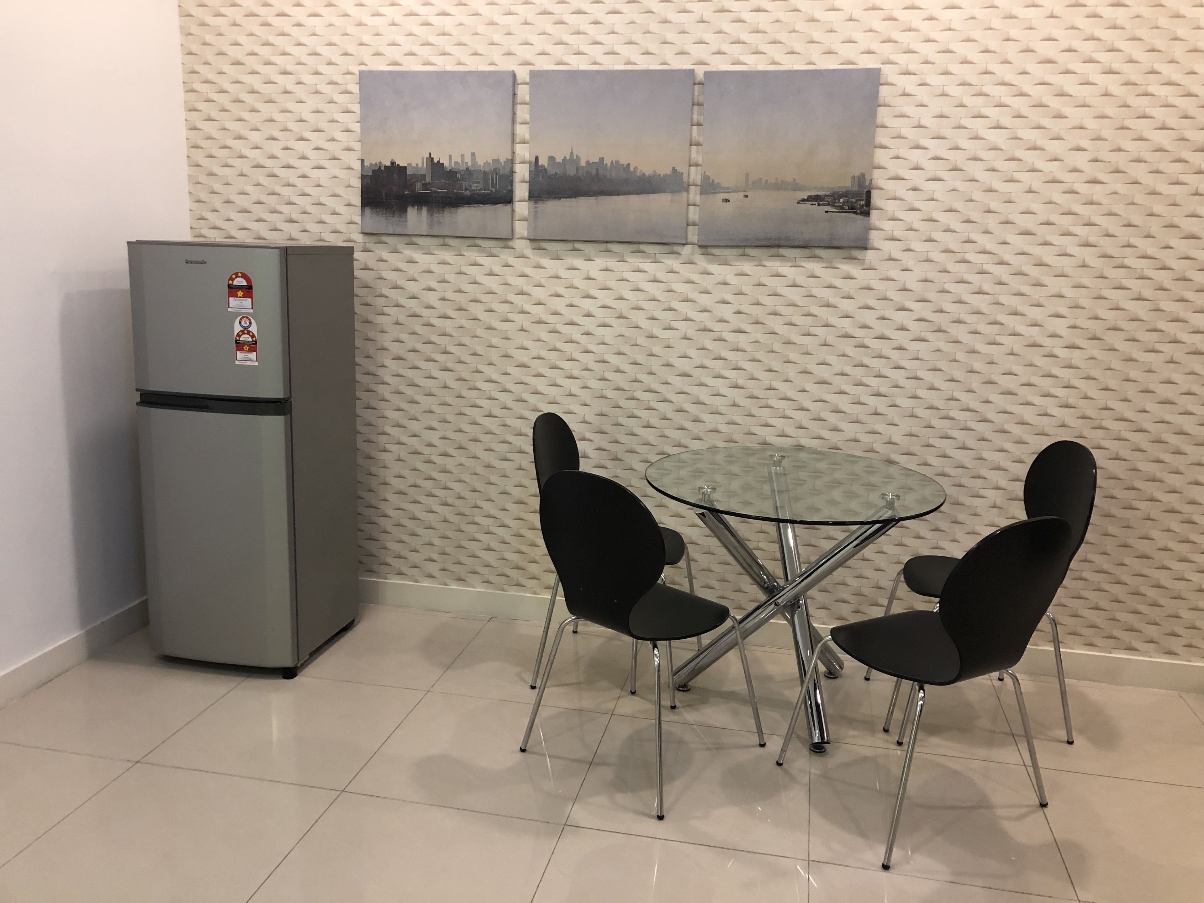 Una habitación pequeña con un refrigerador, dos sillas y una mesa de vidrio. Hay dos cuadros en la pared.