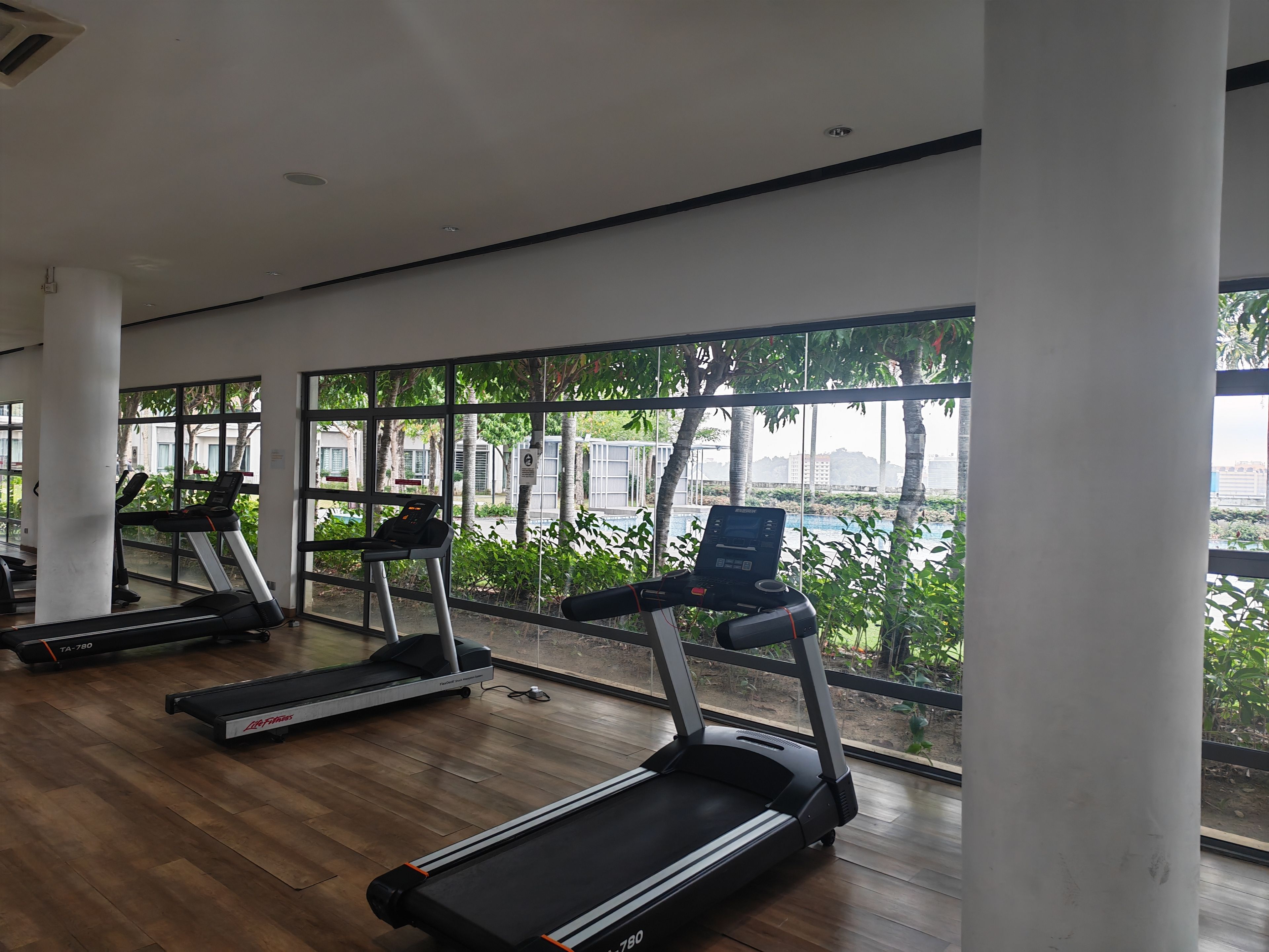 Ein Fitnessraum mit Laufbändern und einem Ergometer, großen Fenstern und Blick auf Grünflächen.