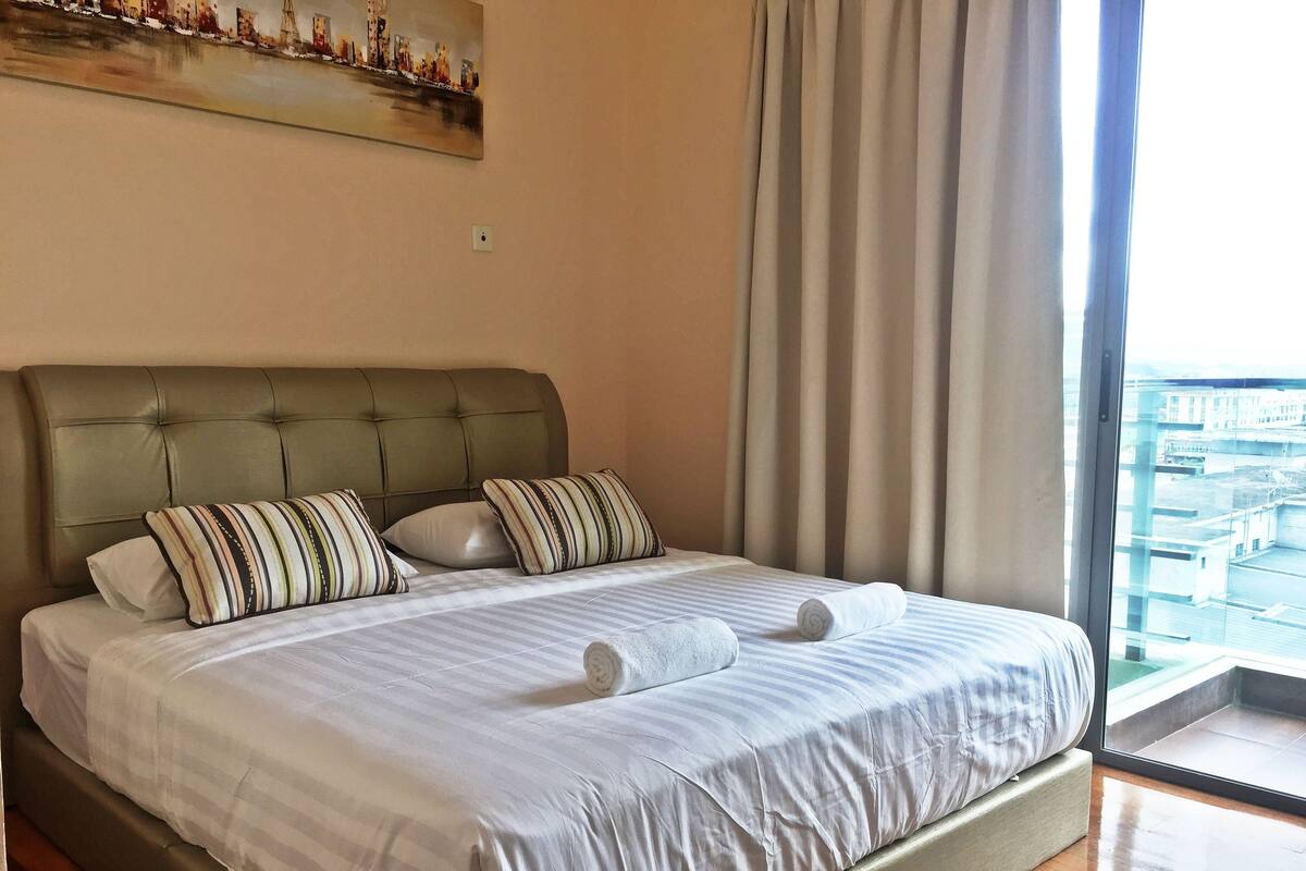 Una cama bien hecha con almohadillas a rayas y un cabecero, un cuadro en la pared y vistas a través de una ventana.