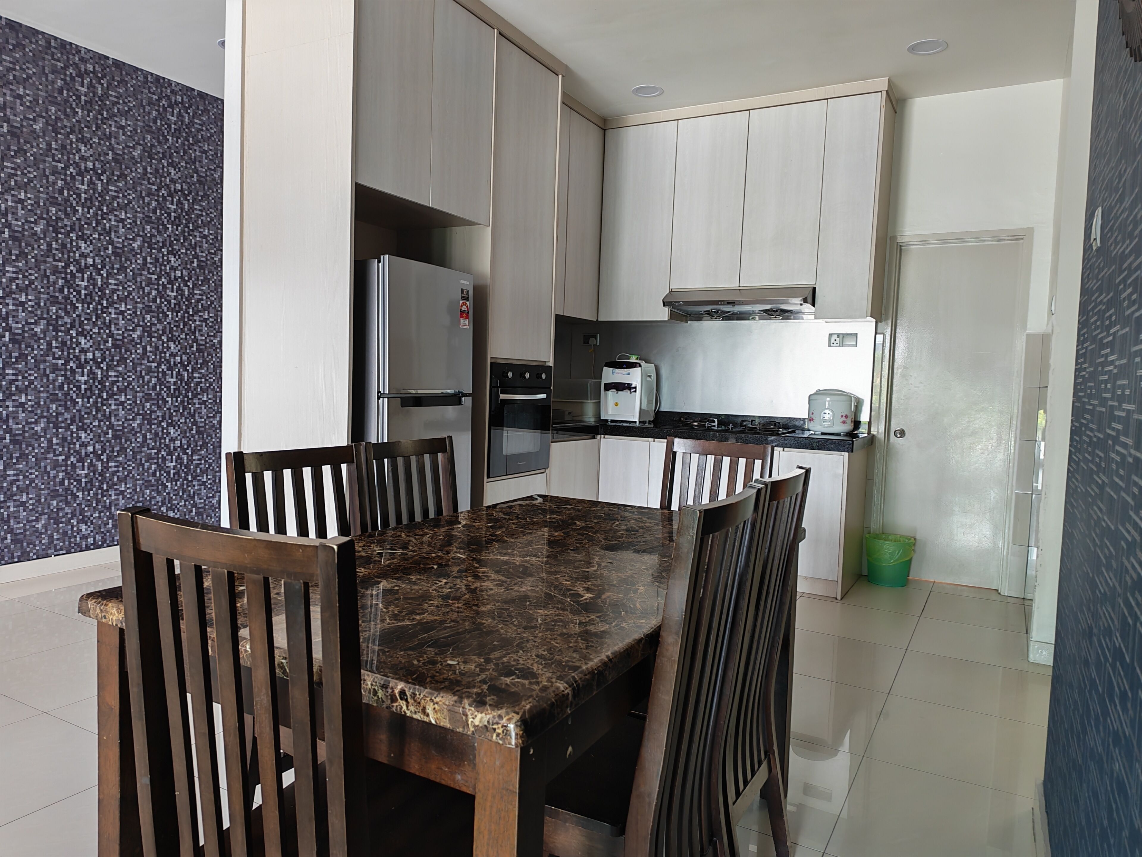 A compact kitchen with a marble countertop, wooden chairs, a built-in oven, and a stainless steel refrigerator.