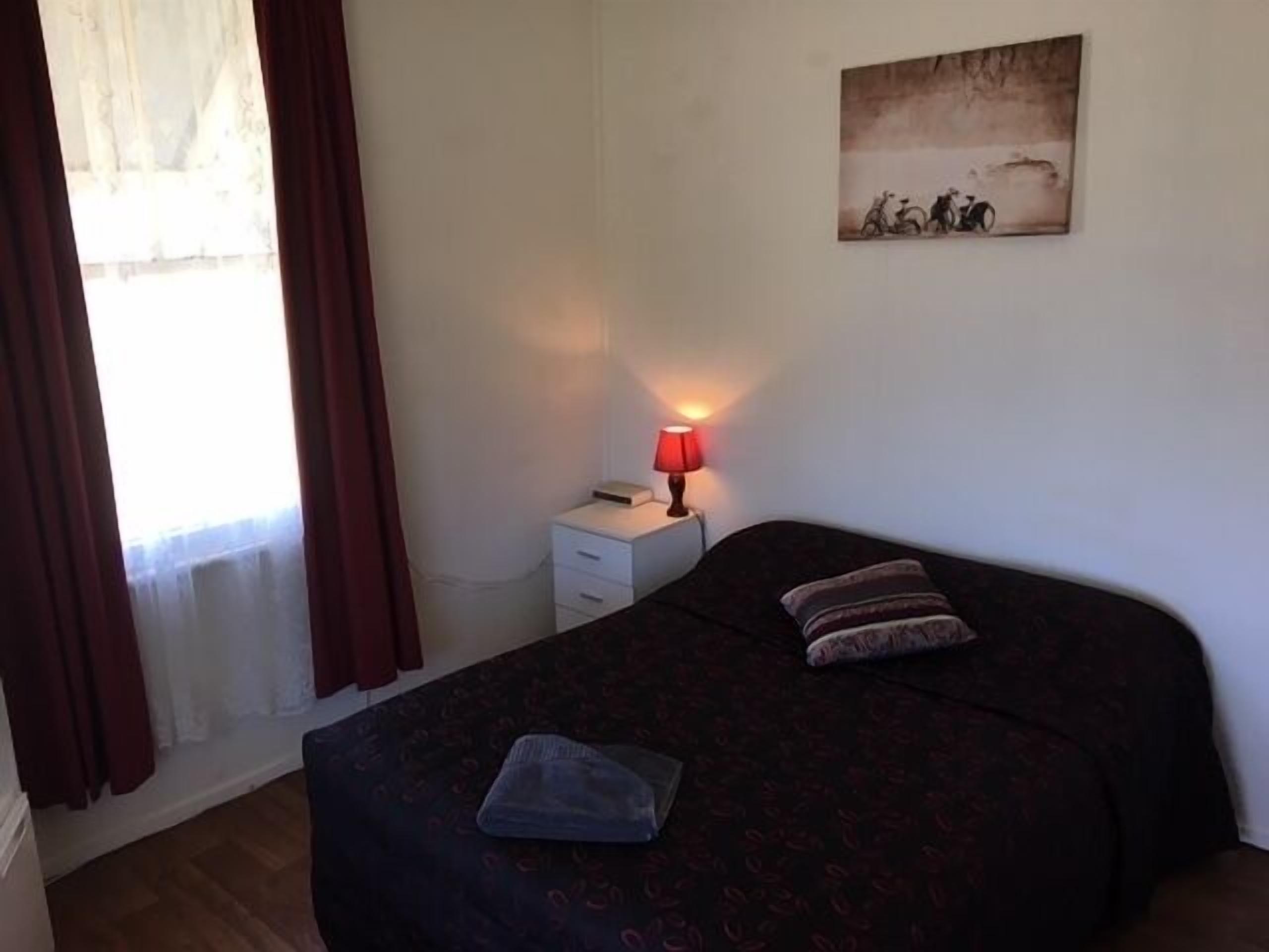 A bedroom with a bed, a nightstand, a lamp, a window with curtains, and a wall-mounted picture.