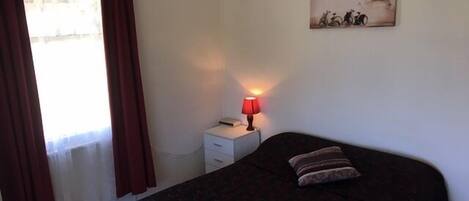 A bedroom with a bed, a nightstand, a lamp, a window with curtains, and a wall-mounted picture.