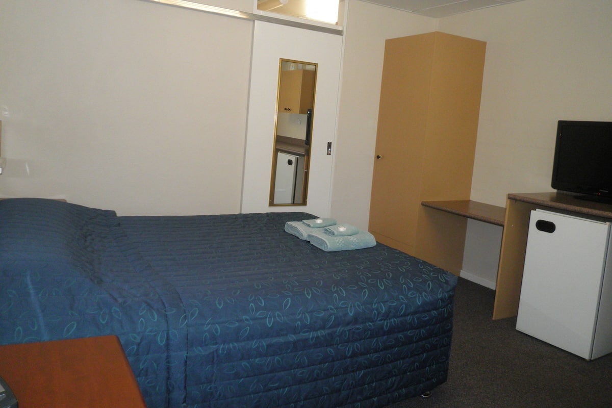 Quarto de hotel com uma cama grande, uma escrivaninha pequena com uma televisão e uma porta que dá para um banheiro.