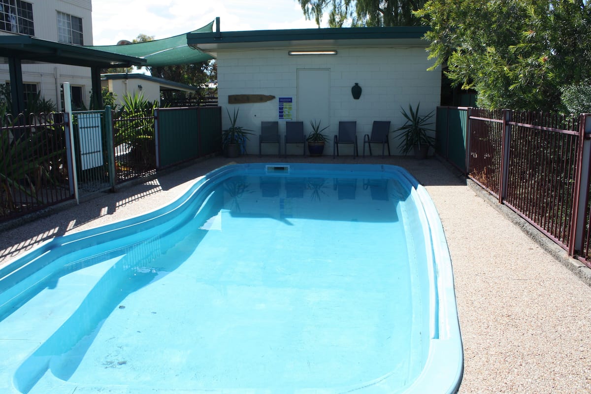 Uma piscina com um pavilhão de telhado verde, um edifício branco com uma porta e um cercado com cadeiras e plantas em vasos.