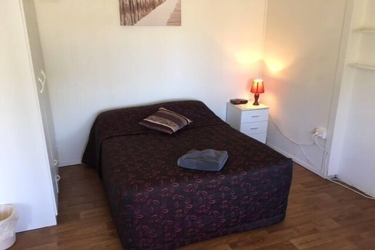 Quarto com cama, mesa de cabeceira com abajur e quadro pendurado na parede.