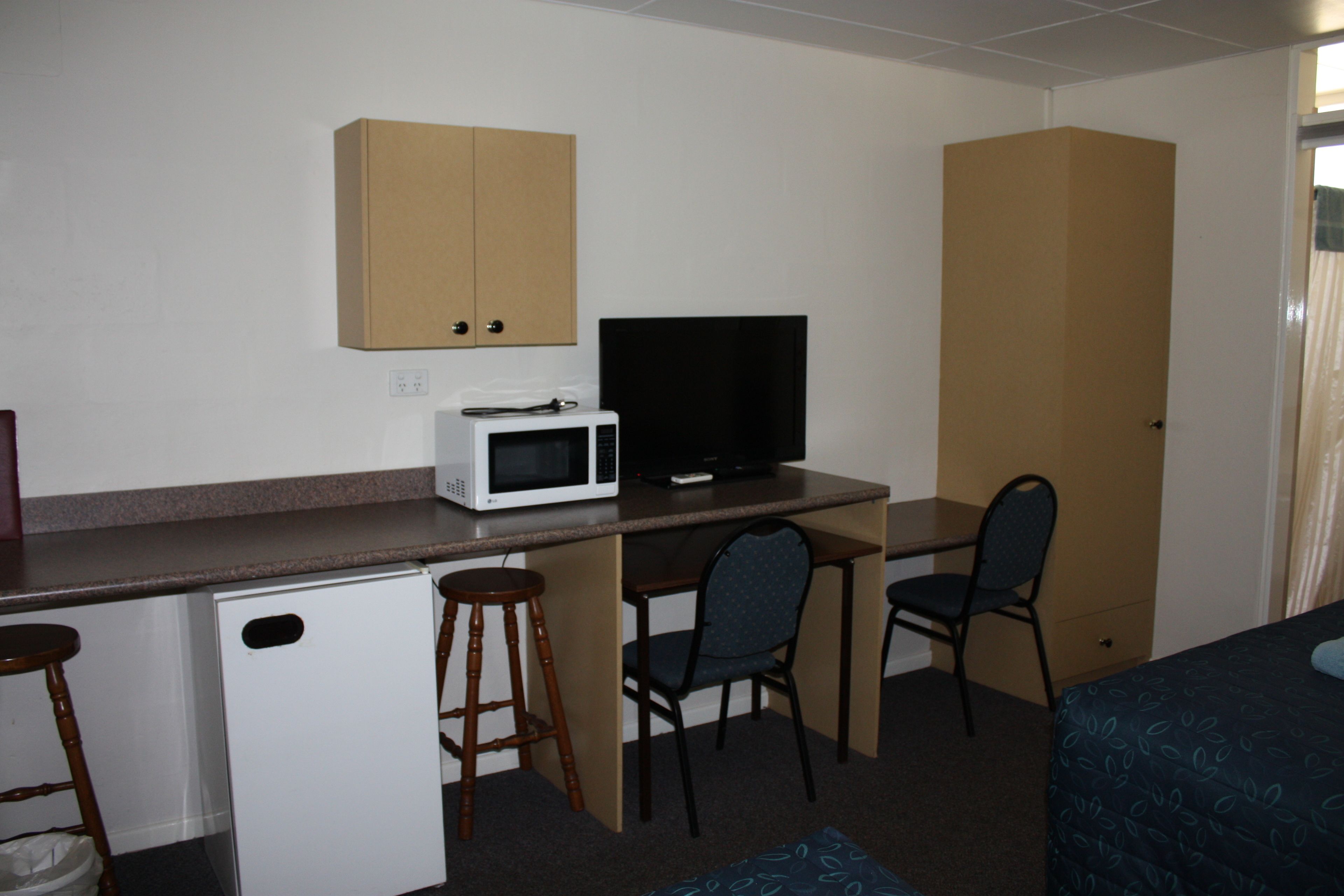 A compact hotel room with a kitchenette, a bed, a desk with a computer and a TV, and two chairs.