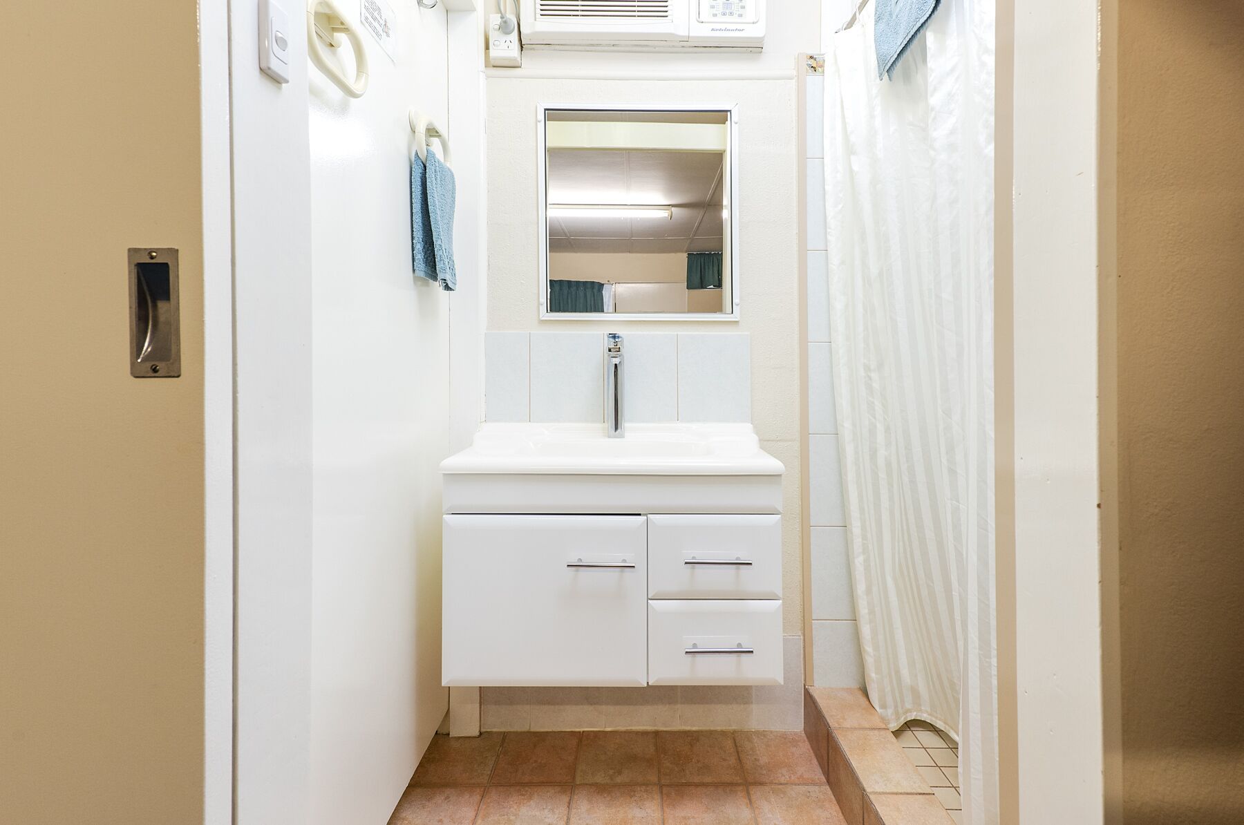 Un pasillo estrecho con un baño blanco que incluye un lavamanos, espejo y toallero.