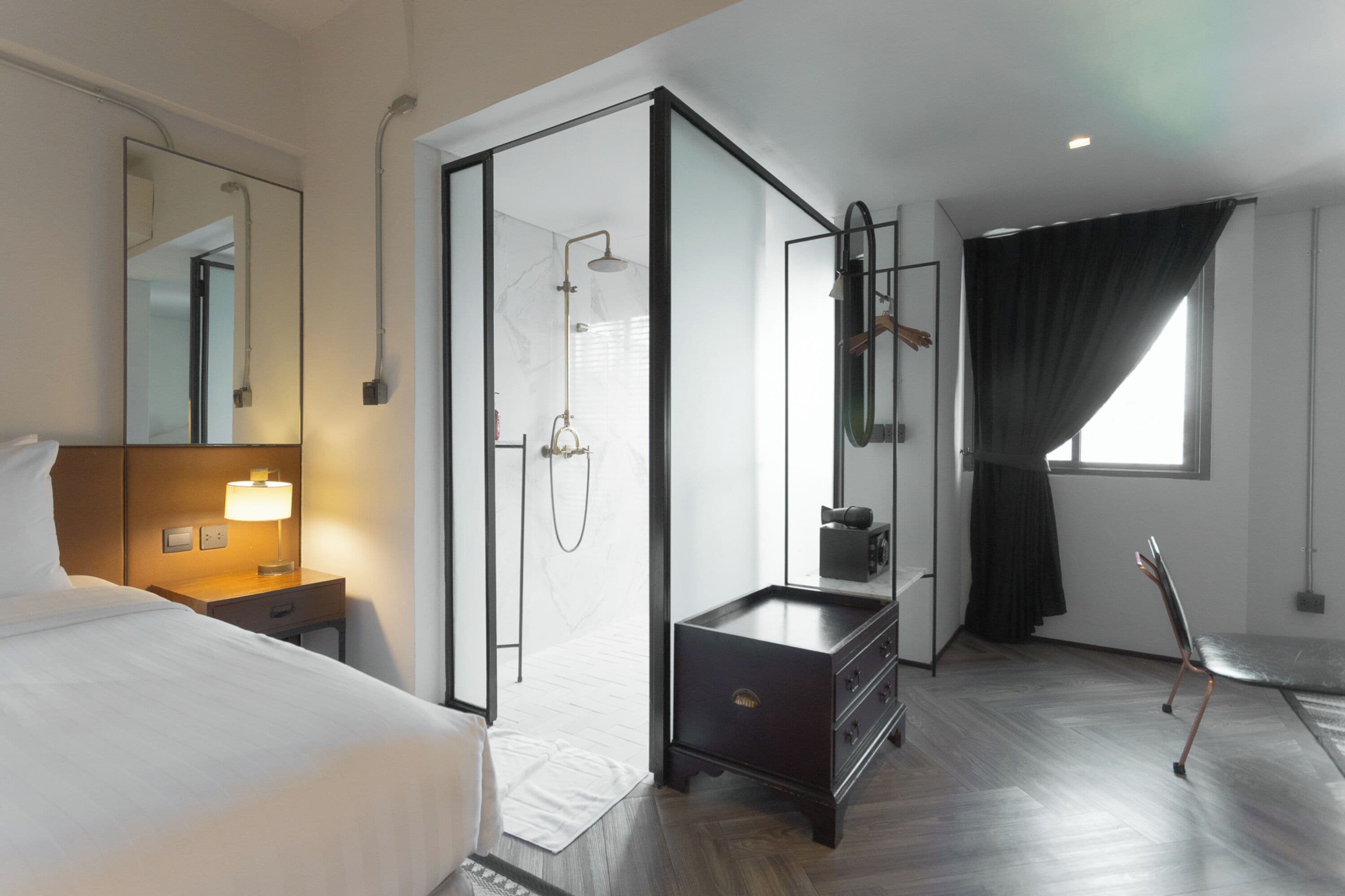 A modern hotel room with a bed, a bedside table with a lamp, a dark wooden dresser, a glass-enclosed shower, and a window with curtains.