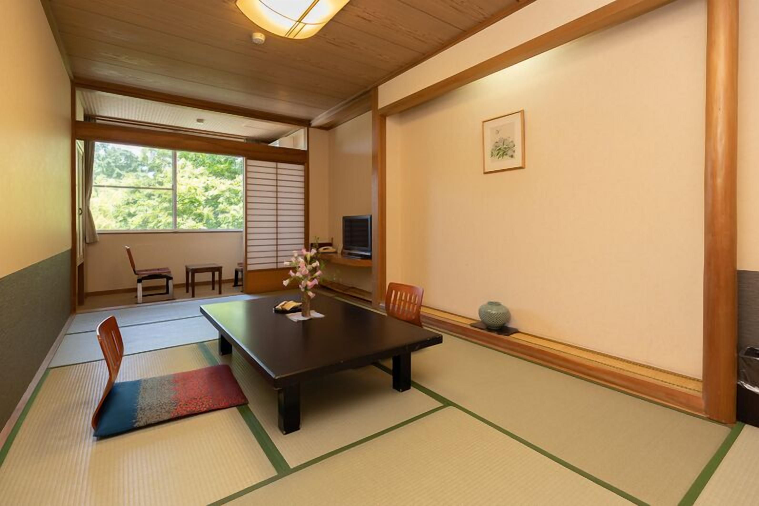 Une pièce de style japonais traditionnel, avec un sol recouvert de tatamis, une table basse, des chaises, un téléviseur et une fenêtre donnant sur un paysage verdoyant.