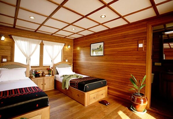 A wooden-paneled bedroom with two beds, a nightstand, a bedside table, a potted plant, and a framed picture on the wall. - Amara River Cruise (Mandalay)