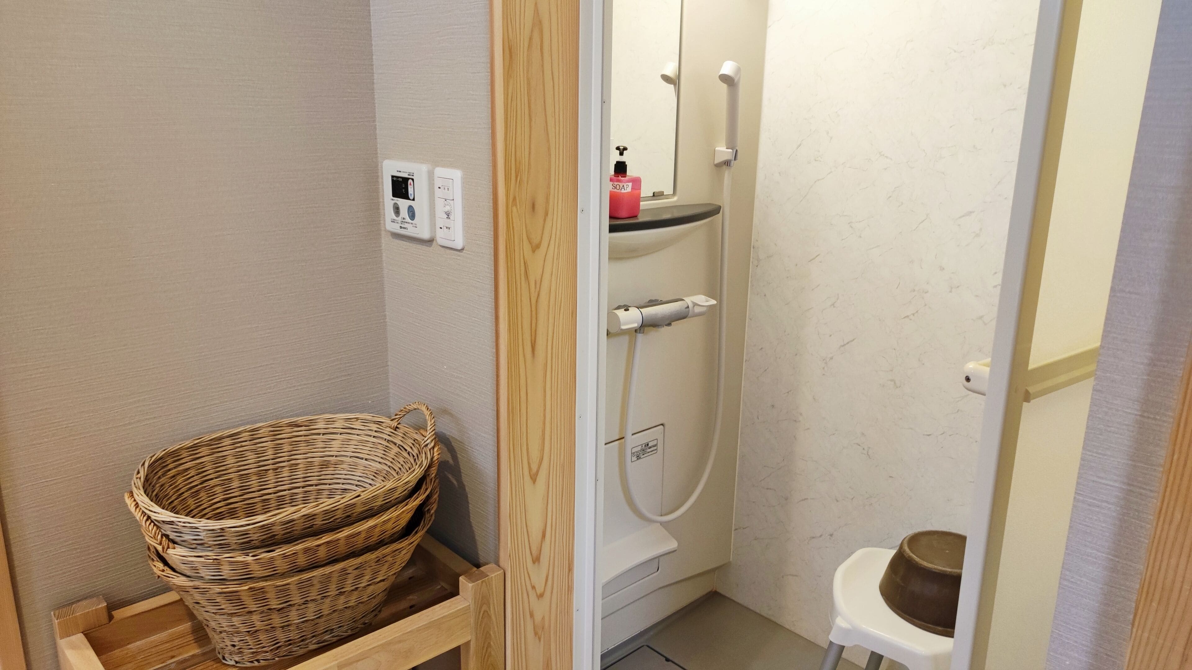 A bathroom with a wooden shelf holding a basket, a white toilet, a shower with a soap dispenser, and a small stool.