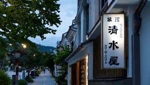 Un edificio japonés tradicional con señalización en japonés, un sendero bordeado de follaje y una vista de la montaña al fondo.