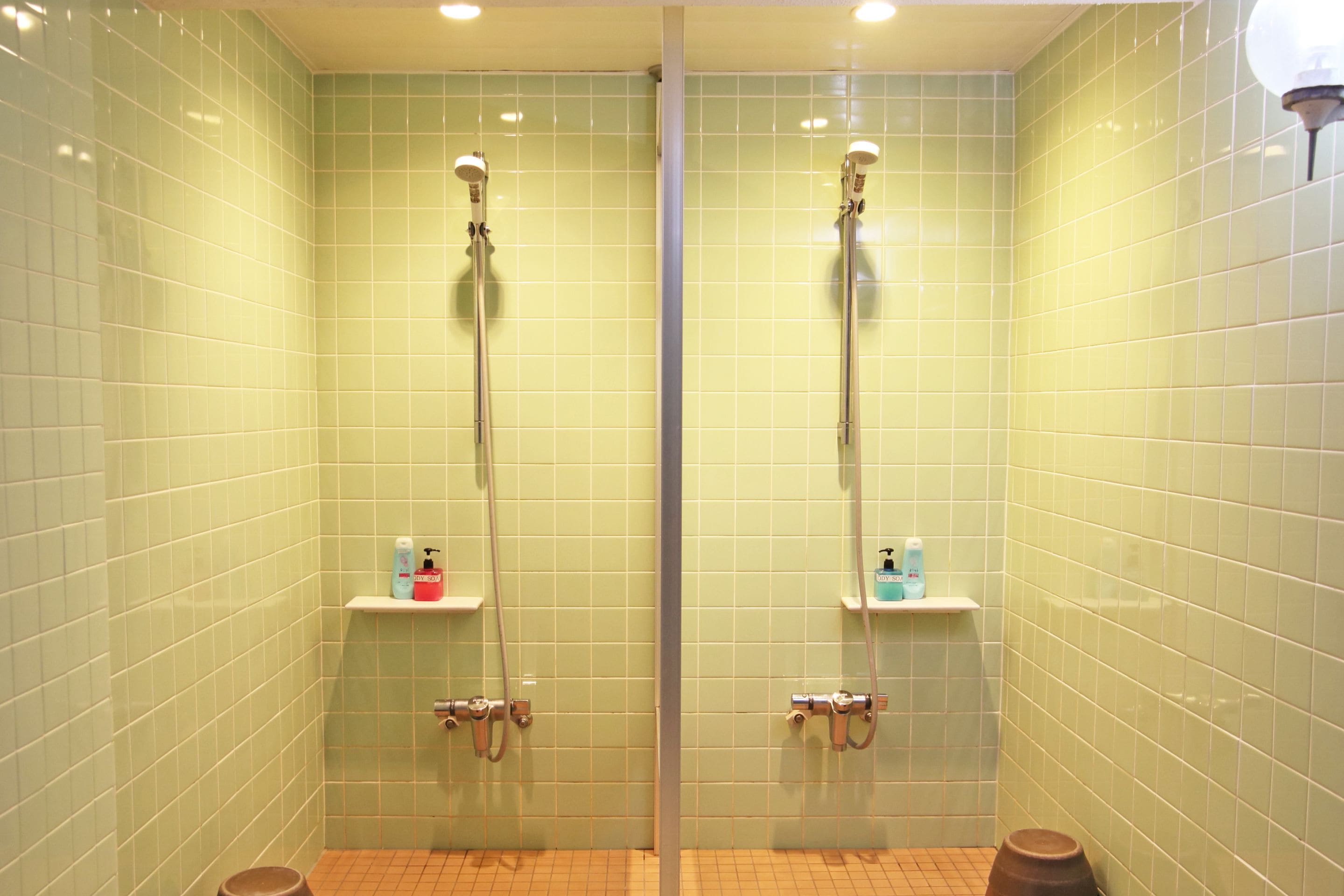 A bathroom with green tiled walls, two showerheads, and shelves with toiletries.