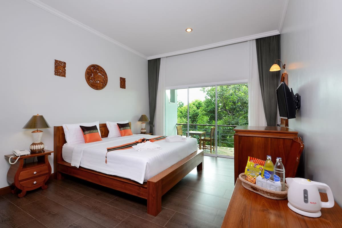Habitación de hotel con una cama grande, mesitas de noche de madera, un escritorio con tetera, un televisor y un balcón con vistas a zonas verdes.