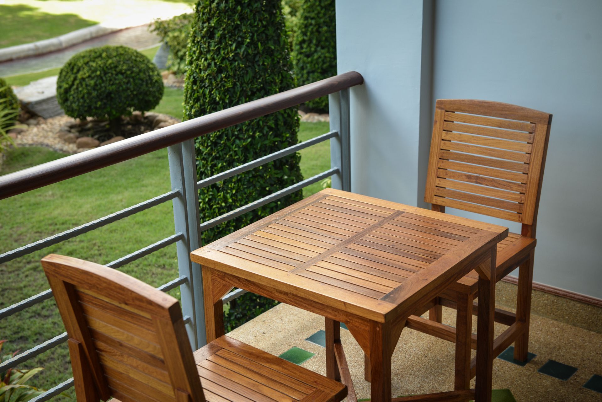 Una mesa de madera y dos sillas en un balcón con baranda de metal, con vista a un jardín con arbustos bien cuidados y césped.