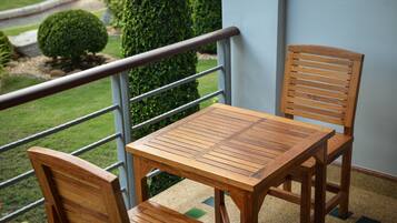 Una mesa de madera y dos sillas en un balcón con baranda de metal, con vista a un jardín con arbustos bien cuidados y césped.