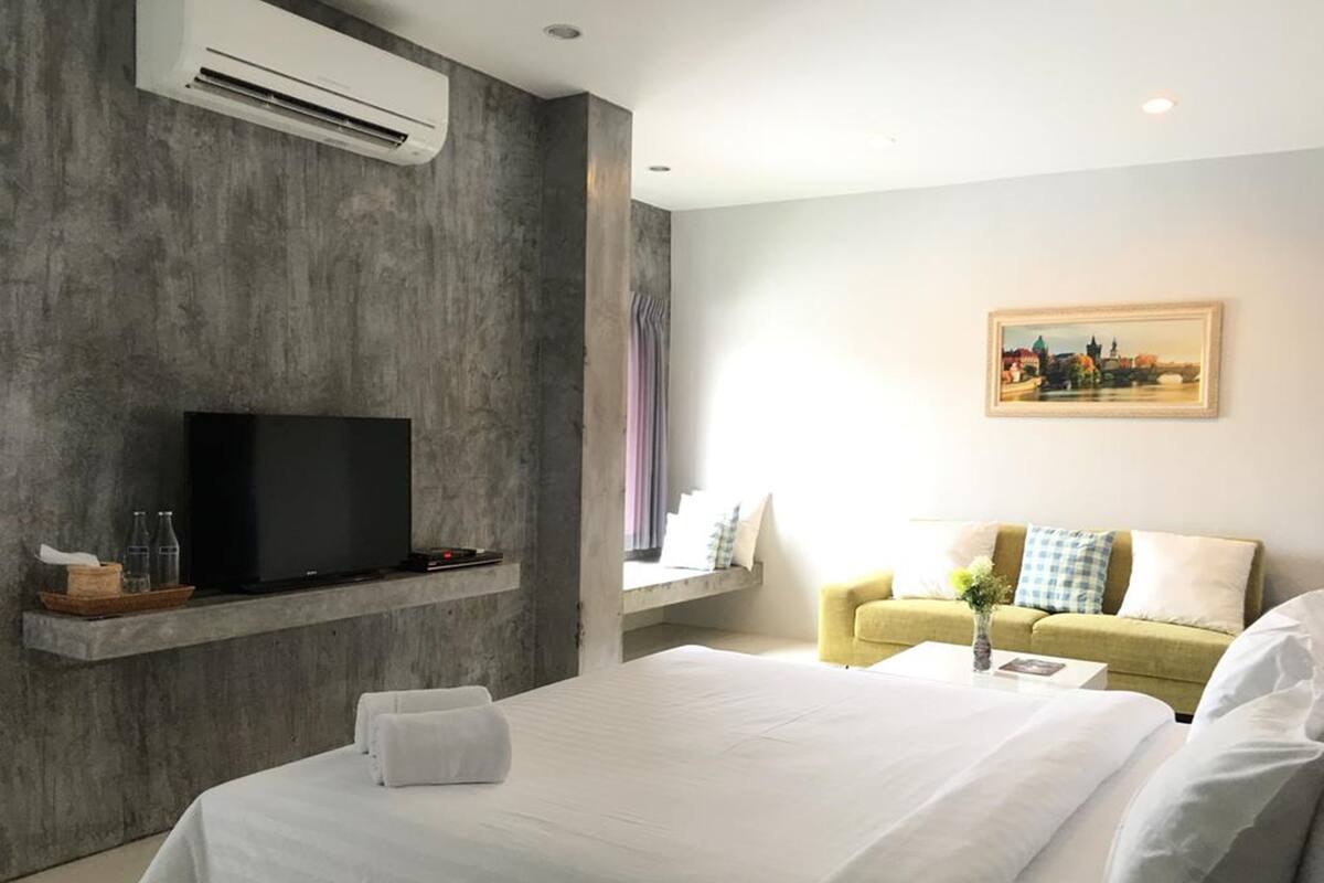Una habitación de hotel moderna con una cama grande, un televisor de pantalla plana en una pared de concreto, un sofá y un cuadro en la pared.